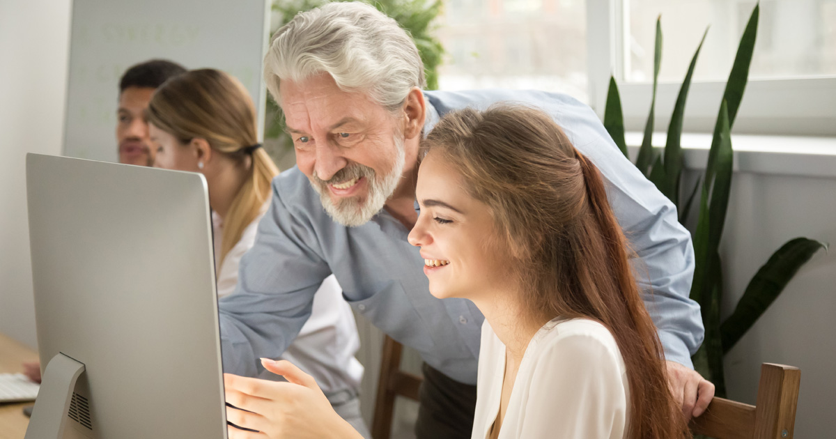 Smiling senior mentor supporting young intern showing achievement result online on computer screen