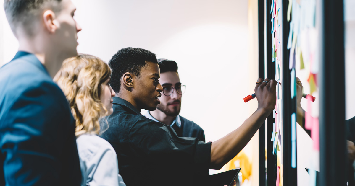 Pensive diverse group of young people dressed in formal wear writing notes on colorful stickers glued on wall during collaborative process in office