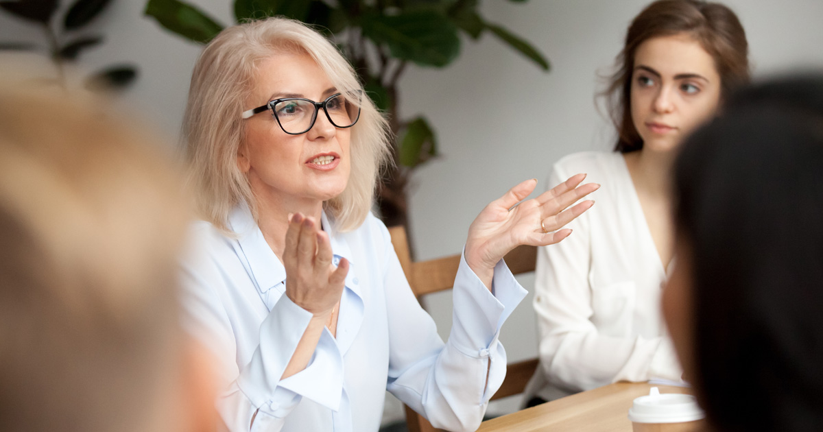 Attractive aged businesswoman speaking to young people, senior woman in glasses teaching audience at training seminar
