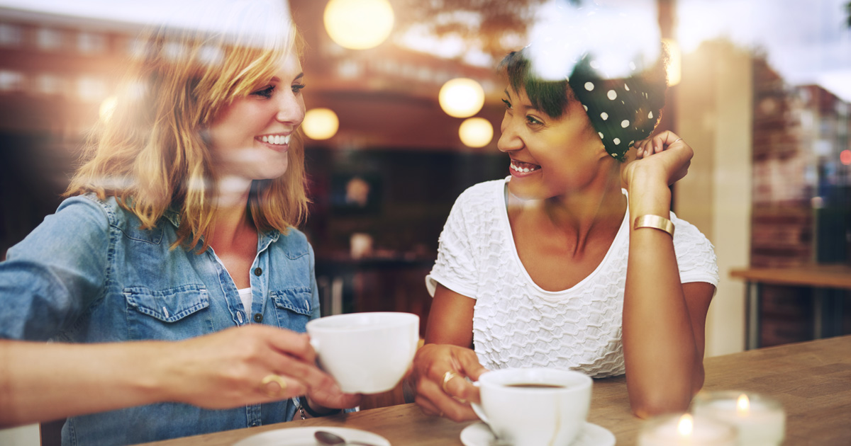 Two friends enjoying coffee together in a coffee shop viewed through glass with reflections as they sit at a table chatting and laughing