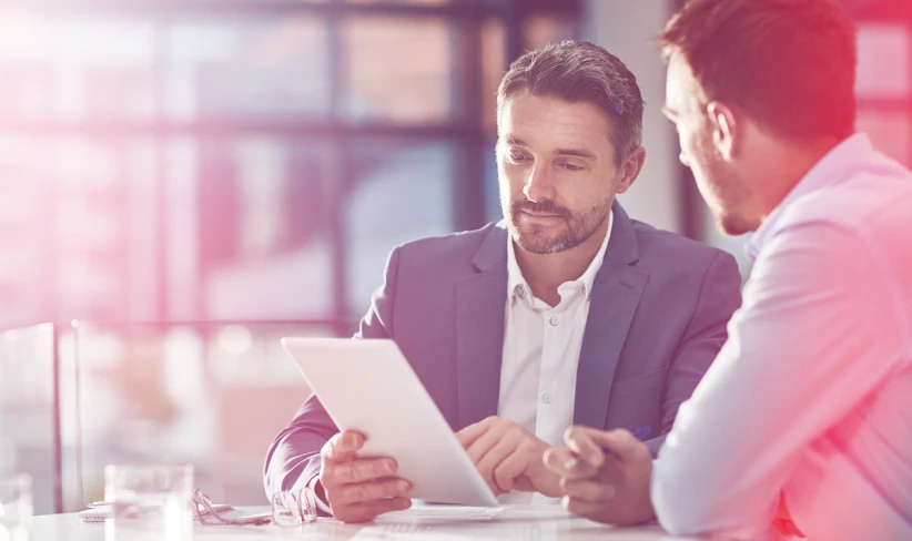 Two men working in an office talking to one another and looking at a tablet.