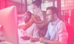 Three people sitting at a desktop, one is pointing at screen while other two take notes.