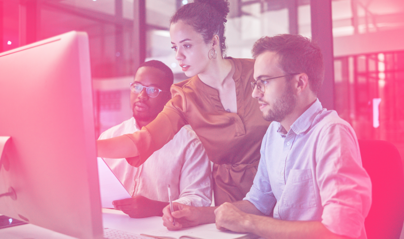 Three people sitting at a desktop, one is pointing at screen while other two take notes.