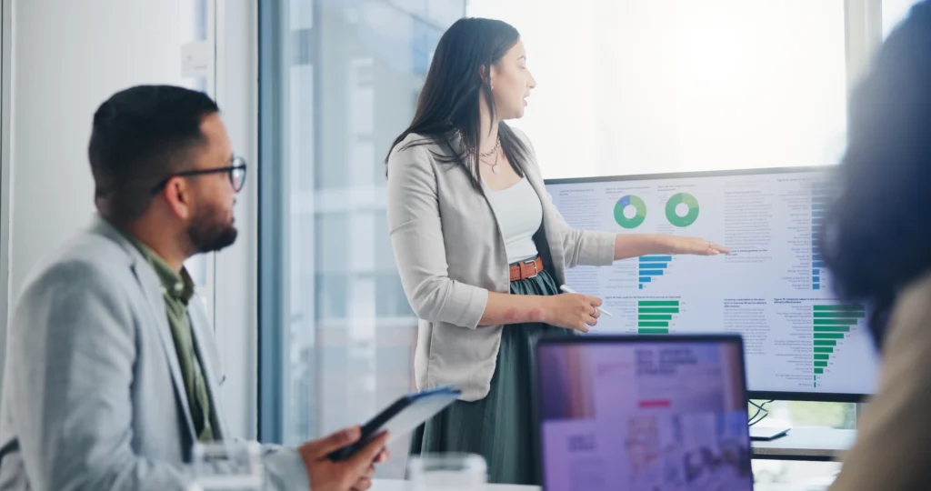 Business professional breaks down data analytics while pointing to a graphical display on a big screen.