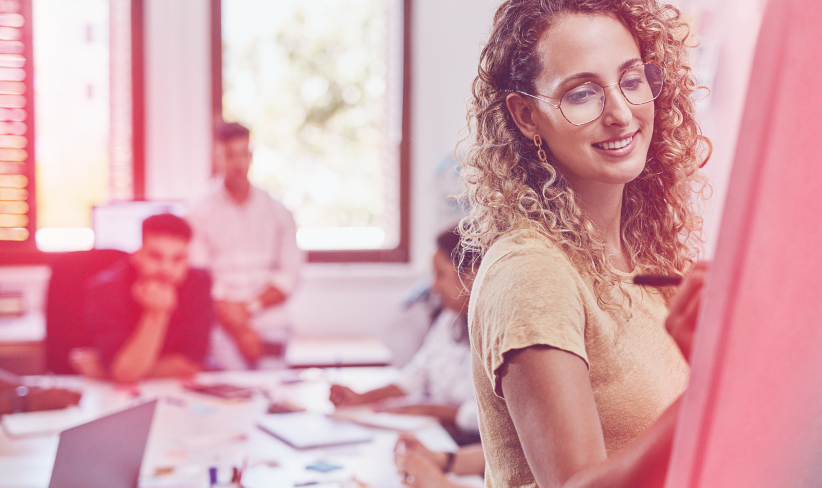 Female office worker writes on whiteboard while colleagues in the background collaborate