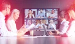 Group of business partners speaking in person and over video call to discuss process improvement plans.